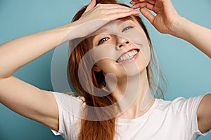 Red-haired girl stands on a blue background in a white T-shirt, smiling, holding a palm on her forehead.