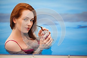 The red-haired girl in a swimsuit standing in a pool. In the hands holding a glass of cold orange juice.