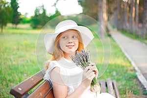 Red-haired girl in a white hat sits on a park bench with a bouquet of lavender