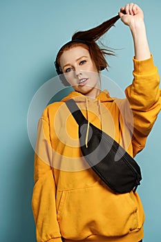 Red-haired girl in a yellow sweatshirt, headphones with a bag over his shoulder stands on a blue background, lifting his