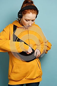 Red-haired girl in a yellow sweatshirt and headphones stands on a blue background, buttoning a bag over shoulders with