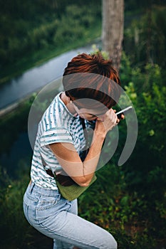 Red-haired hipster girl with a phone, stands against the background of the river. Processing under the film.
