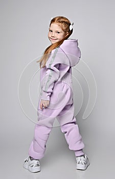 Red-haired kid girl in pink jumpsuit with hood stands back to camera with legs wide apart and turned to camera