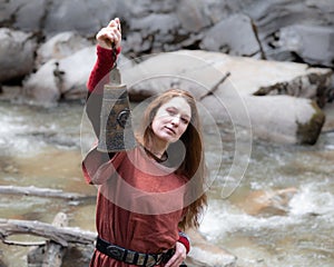 Red-haired long-haired woman holding an antique bell high in her hands against the backdrop of a mountain river, Austria