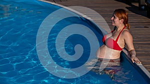 A red-haired pregnant woman in a red bikini is resting in the pool, leaning on the side.