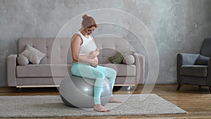 Red-haired pregnant woman stroking her belly while sitting on a fitball at home.