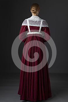 A Regency maid servant wearing a red linen dress with an apron and a lace modesty shawl against a studio backdrop photo