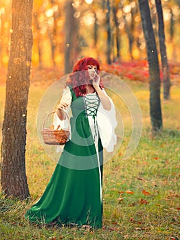 Red-haired superb princess walks across the field and gathers herbs for sorceress`s magic potion, dressed in green dress