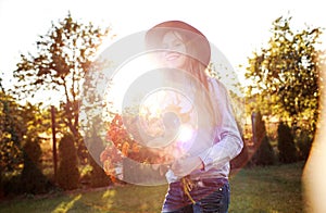 a red-haired teenage girl in a red hat dances on the lawn with a bouquet of flowers against the sunset background