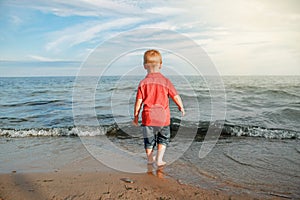 Toddler child kid in red t-shirt and jeans standing near water on lake sea ocean beach at evening sunset looking far away.