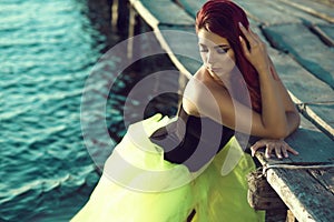 Red haired woman in black corset and long tail green veiling skirt standing in the sea water leaning on the pier. Mermaid concept