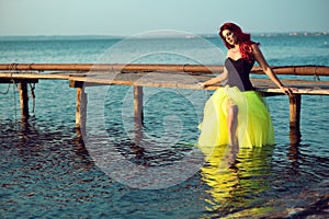 Red haired woman in black corset and long tail green veiling skirt standing in sea water and leaning on wooden pier.