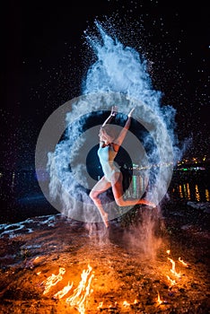 Red-haired woman in a blue swimsuit dancing in clouds of flour in front of a fire. Girl jumping scattering white powder