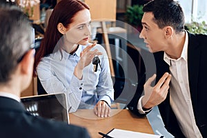 Red-haired woman is holding on finger keys sitting next to adult man in lawyer`s office.