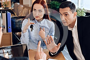 Red-haired woman is holding on finger keys sitting next to adult man in lawyer`s office.