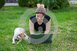 A red-haired woman lies on the green grass in a park with her own dog. The girl maintains a social distance and types on