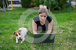 A red-haired woman lies on the green grass in a park with her own dog. The girl maintains a social distance and types on