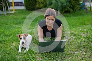 A red-haired woman lies on the green grass in a park with her own dog. The girl maintains a social distance and types on photo