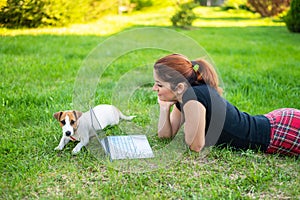 The red-haired woman lies on a green grass in a park with a puppy. The girl maintains a social distance and watches a