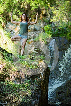 Red-haired woman in shorts doing yoga at the waterfall. A girl stands in a pose of a tree in the forest