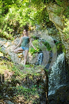 Red-haired woman in shorts doing yoga at the waterfall. A girl stands in a pose of a tree in the forest