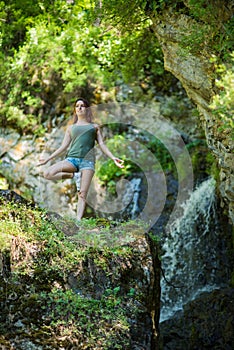 Red-haired woman in shorts doing yoga at the waterfall. A girl stands in a pose of a tree in the forest