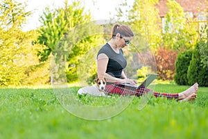 Red-haired woman sits in green grass with a puppy in the park and online shopping. The girl maintains a social distance
