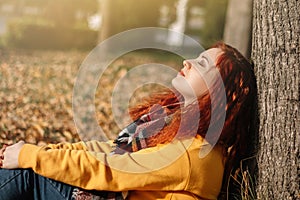 Red-haired woman sitting on ground leaning against tree and enjoying sunny autumn day.