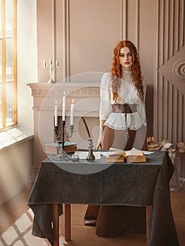 Red-haired woman in vintage dress stands in classic room, window sun light. Clothing costume countess old style white