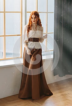 Red-haired woman in vintage dress stands at large classic window waiting love. Clothing costume countess old style white