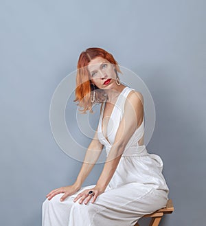 Red-haired woman in white sitting on a wooden chair. Looks into the camera.. Red lipstick. Deep neckline. Hands on knees