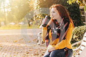 Red-haired woman in yellow sweatshirt sits on park bench and drinks coffee. She is holding reusable mug in her hands