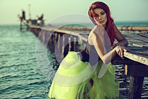 Red haired womanstanding in the sea water leaning on the pier. Mermaid coming out of water and trying to make first steps