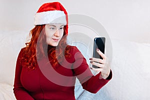 Red-haired young woman in Santa hat uses video chat on her mobile phone. Online greeting with Christmas