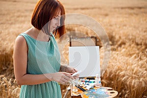 Red-headed girl in light blue dress painting a picture in wheat field