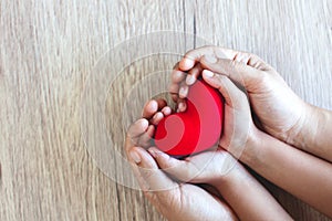 Red heart in child hands and parent hands on wooden table background