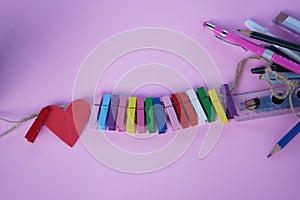 Red heart with colorful wooden clips on rope and pencil pens on soft pink and purple background. Creative concept