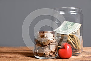 Red heart and donation jars with money on wooden table against grey background.