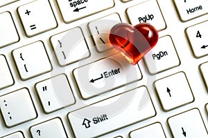 A red heart lies on a white keyboard for a computer next to the enter key close-up. The concept of likes for social networks,