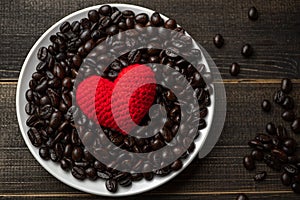 Red heart on a lot of roasted coffee beans background on white plate on wooden table. Strong black espresso, Grains of coffee