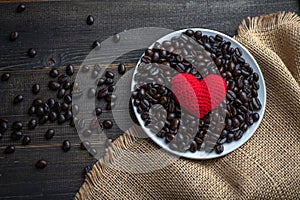 Red heart on a lot of roasted coffee beans background on white plate on wooden table. Strong black espresso, Grains of coffee back