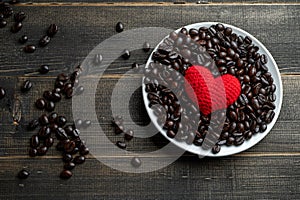 Red heart on a lot of roasted coffee beans background on white plate on wooden table. Strong black espresso, Grains of coffee back