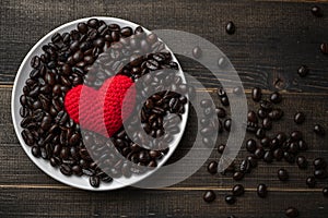 Red heart on a lot of roasted coffee beans background on white plate on wooden table. Strong black espresso, Grains of coffee back