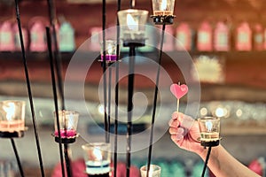 A red heart shaped candle in a woman`s hand with candlelight in many small glass cups.