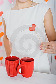 Red heart on a stick in a female hand on a white dress next to two red cups.