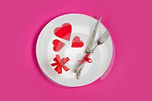 Red hearts on a white plate  a fork  a knife  a gift box on a pink background  top view  copy space. Valentine`s day