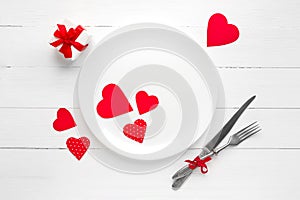 Red hearts on a white plate  a fork  a knife  a red ribbon  a gift box on a white wooden table  top view  copy space. Wedding