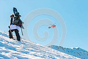 Red helicopter flying over snowy mountains and a snowboarder is hiking