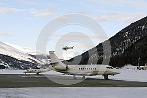 A red helicopter flying over two private jets in the airport of St Moritz Switzerland in winter