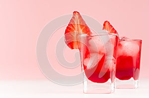 Red homemade liquors in two shot glasses with ice cubes, strawberry slice on soft pastel pink background and white wood table.